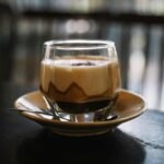 Close-up of a glass cup of foamy cappuccino with saucer and teaspoon on a table.